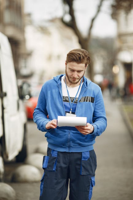 A young man with light brown hair and a beard, wearing a blue hoodie with black accents and dark blue work trousers with multiple pockets, stands outdoors on a street in Fulham. He is holding and read