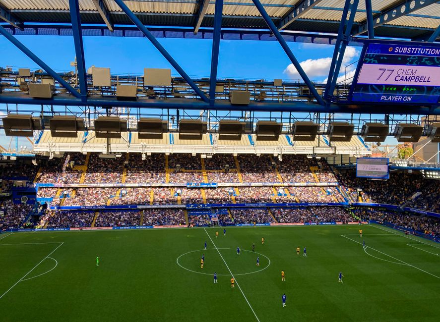 A large modern stadium with a distinctive architectural design featuring a white, geometric framework supporting the roof structure, situated outdoors under a clear sky. In the foreground, a pedestria